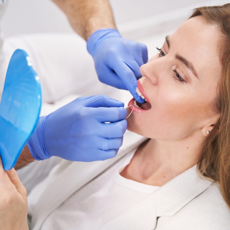 Female patient lying in dental chair and looking in the mirror while doctor showing how to use dental floss
