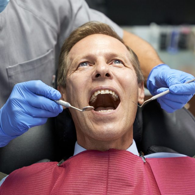 Fair-haired man opening mouth for dentist to check his teeth with pick and mirror