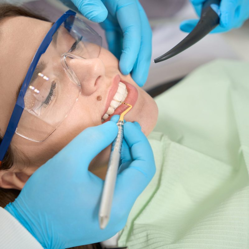 Female client in protective eyewear lying supine while professional dentist removing tartar between her teeth